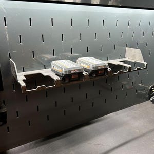 Może przedstawiać: Czarna metalowa tablica perforowana z p&oacute;łką, na kt&oacute;rej znajdują się dwa szare i czarne akumulatory Ridgid 8.0 Ah. Tablica perforowana ma r&oacute;wnomiernie rozmieszczone szczeliny do organizacji narzędzi. P&oacute;łka jest zamontowana na tablicy.