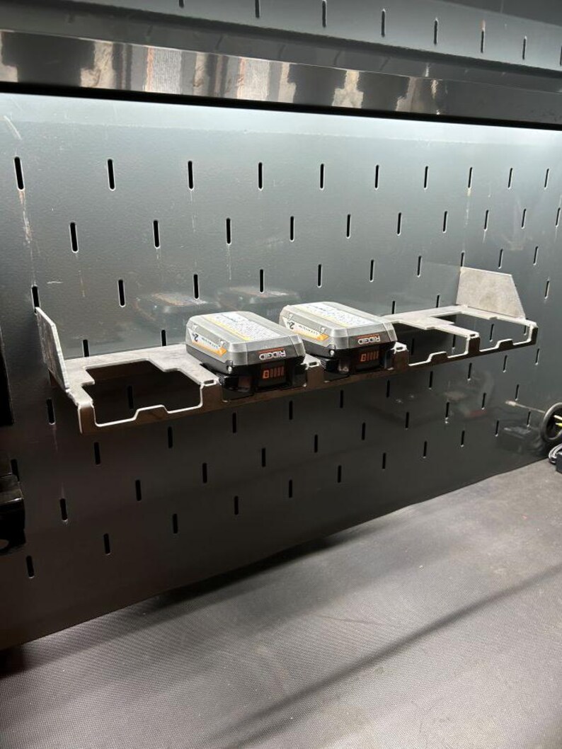 Może przedstawiać: Czarna metalowa tablica perforowana z p&oacute;łką, na kt&oacute;rej znajdują się dwa szare i czarne akumulatory Ridgid 8.0 Ah. Tablica perforowana ma r&oacute;wnomiernie rozmieszczone szczeliny do organizacji narzędzi. P&oacute;łka jest zamontowana na tablicy.