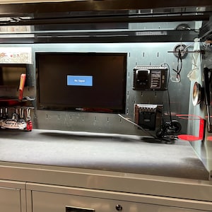 May include: A gray metal workbench with a black flat-screen TV displaying "No Signal." The workbench has drawers, a pegboard, and various tools and accessories. Red and black accessories are visible.