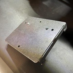 Może przedstawiać: Srebrna metalowa płyta z kilkoma otworami i zagiętą krawędzią. Widoczne są dwie śruby mocujące płytę. Płyta ma prostokątny kształt ze ściętym rogiem.