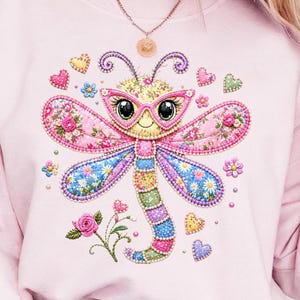 May include: A light pink sweatshirt featuring a whimsical dragonfly design. The dragonfly has pink floral wings, a colorful body, and wears pink glasses. Decorative hearts and flowers surround the dragonfly, creating a cheerful and artistic design.