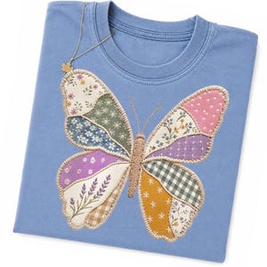 Può includere: Una t-shirt blu pervinca con una grande applicazione a forma di farfalla sul davanti. Le ali della farfalla sono composte da vari ritagli di tessuto con motivi floreali, a quadretti e a pois nelle tonalità del viola, verde, giallo e bianco.