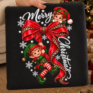 Peut inclure: Un sweat-shirt noir avec un motif représentant deux lutins en tenues vertes et rouges, un grand nœud rouge à motif léopard et des flocons de neige blancs. Le texte Merry Christmas est écrit en lettres blanches sur le motif.