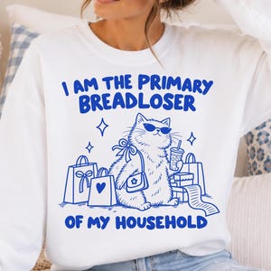 May include: A white crewneck sweatshirt featuring a blue graphic of a cat wearing sunglasses, carrying a purse, and surrounded by shopping bags. The text reads, I AM THE PRIMARY BREADLOSER OF MY HOUSEHOLD, printed in a bold blue sans-serif font.