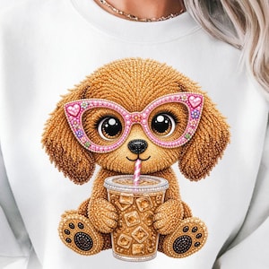 May include: A white sweatshirt with a cartoon dog wearing pink glasses with heart-shaped details. The dog is holding a cup of iced coffee with a pink straw. The dog is brown with large, expressive eyes.