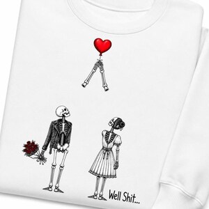 May include: A white sweatshirt featuring a graphic of two skeletons. One skeleton wears a leather jacket and holds a bouquet of red roses, while the other wears a dress. Above them, a pair of skeletal hands holds a red heart-shaped balloon. The text Well Shit... is printed in black near the bottom.