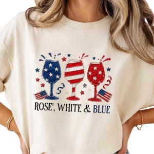 Może przedstawiać: Kremowa koszulka z trzema kieliszkami do wina w patriotycznych wzorach w kolorach czerwonym, białym i niebieskim. Pod spodem nadruk "ROSE', WHITE & BLUE", a także małe amerykańskie flagi i gwiazdy.