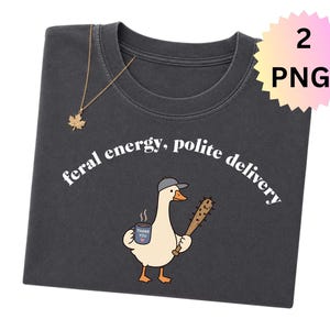 Pode incluir: Camiseta cinza escuro com a frase "feral energy, polite delivery" em fonte branca curva. A camiseta apresenta um ganso de desenho animado com um chapéu, segurando um bastão e uma caneca. Um colar dourado com pingente de folha de bordo está sobre a camiseta.