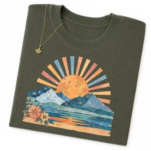 May include: Olive green crew neck t-shirt with a colorful mountain and sun graphic. The design features a sun with orange, blue, and pink rays, mountains, and flowers. A gold leaf pendant hangs on the shirt.