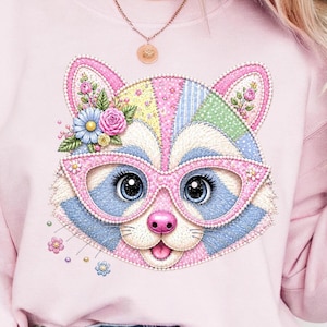 Könnte beinhalten: Hellrosa Sweatshirt mit einem bunten Waschbär-Design. Der Waschbär trägt eine rosa Brille und einen floralen Kopfschmuck. Das Design umfasst blaue, rosa, gelbe und grüne Patches mit einer Perlenkontur. Der Waschbär hat große blaue Augen und eine rosa Nase.
