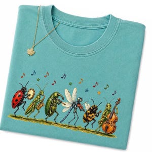 Puede incluir: Una camiseta de cuello redondo color verde azulado con un gráfico de insectos tocando instrumentos musicales, incluyendo una mariquita, saltamontes, escarabajo, libélula, abeja y grillo. Un collar con dije de hoja de arce dorado descansa arriba.