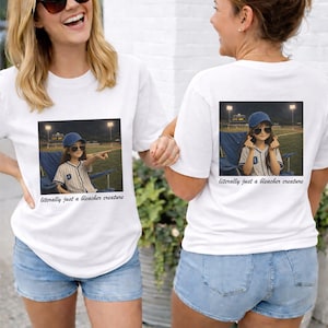 Puede incluir: Camisetas blancas con un gráfico de un niño con uniforme de béisbol, con gafas de sol y gorra de béisbol, señalando. El texto "literally just a bleacher creature" está debajo de la imagen. Las mujeres llevan pantalones cortos vaqueros.