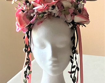 May include: A pink floral crown with antlers, featuring pink roses, white flowers, and green leaves. The crown is adorned with black and pink ribbons.