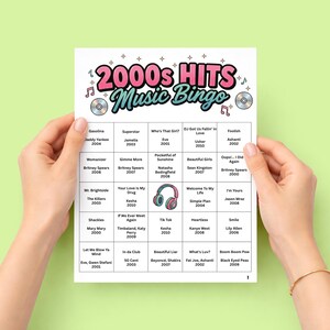 May include: A sheet of 2000s Hits Music Bingo, held by two hands against a light green background. The bingo card features a colorful design with the text "2000s Hits Music Bingo" and a grid of music artists and song titles.