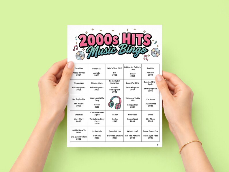May include: A sheet of 2000s Hits Music Bingo, held by two hands against a light green background. The bingo card features a colorful design with the text "2000s Hits Music Bingo" and a grid of music artists and song titles.