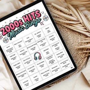 May include: A digital tablet displaying a 2000s Hits Music Bingo game. The game board features song titles and artist names from the 2000s, with a colorful design including musical notes and CD graphics. The tablet is on a neutral-toned surface.