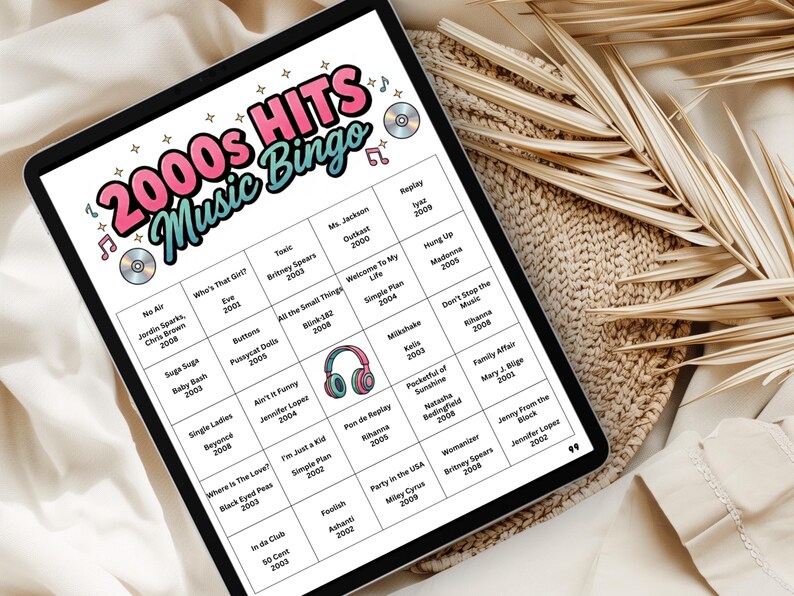 May include: A digital tablet displaying a 2000s Hits Music Bingo game. The game board features song titles and artist names from the 2000s, with a colorful design including musical notes and CD graphics. The tablet is on a neutral-toned surface.