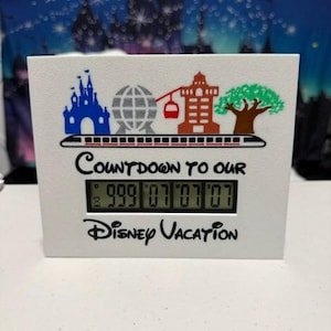 Puede incluir: Un reloj digital blanco con gráficos coloridos de Disney. El reloj muestra "999 07 07 07" y el texto "Countdown to our Disney Vacation". La parte superior presenta ilustraciones de un castillo, un globo terráqueo y un árbol.