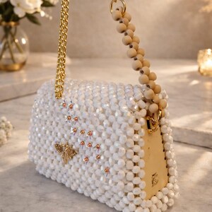 Peut inclure: Sac &agrave; main blanc perl&eacute; avec une cha&icirc;ne dor&eacute;e et une poign&eacute;e en perles de bois. Le sac rectangulaire pr&eacute;sente une libellule dor&eacute;e et de petits accents orange. Le sac est orn&eacute; de perles blanches et poss&egrave;de un fermoir dor&eacute;.