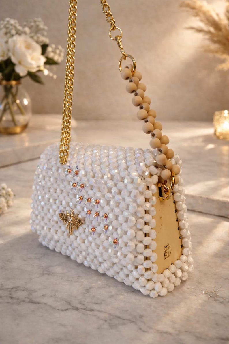 Peut inclure: Sac &agrave; main blanc perl&eacute; avec une cha&icirc;ne dor&eacute;e et une poign&eacute;e en perles de bois. Le sac rectangulaire pr&eacute;sente une libellule dor&eacute;e et de petits accents orange. Le sac est orn&eacute; de perles blanches et poss&egrave;de un fermoir dor&eacute;.