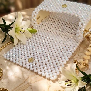 Peut inclure: Un sac &agrave; main blanc perl&eacute; avec une cha&icirc;ne et un fermoir dor&eacute;s. Le sac est ouvert, r&eacute;v&eacute;lant un int&eacute;rieur de couleur claire. Deux fleurs de lys blanches avec des &eacute;tamines jaunes sont plac&eacute;es pr&egrave;s du sac, et une cha&icirc;ne dor&eacute;e est drap&eacute;e autour.