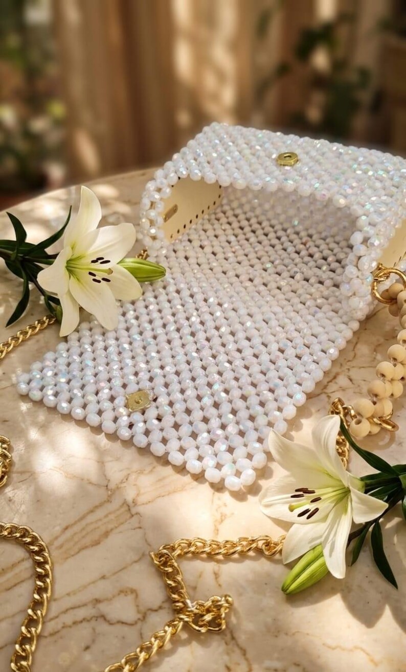 Peut inclure: Un sac &agrave; main blanc perl&eacute; avec une cha&icirc;ne et un fermoir dor&eacute;s. Le sac est ouvert, r&eacute;v&eacute;lant un int&eacute;rieur de couleur claire. Deux fleurs de lys blanches avec des &eacute;tamines jaunes sont plac&eacute;es pr&egrave;s du sac, et une cha&icirc;ne dor&eacute;e est drap&eacute;e autour.
