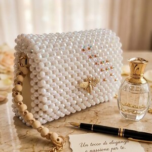 Peut inclure: Un sac &agrave; main blanc perl&eacute; avec une abeille dor&eacute;e et des accents floraux. Le sac a une poign&eacute;e en perles de bois et une bandouli&egrave;re en cha&icirc;ne dor&eacute;e. Un flacon de parfum et un stylo sont &agrave; c&ocirc;t&eacute; du sac. Le texte sur la carte dit "Un tocco di eleganza e passione per te."