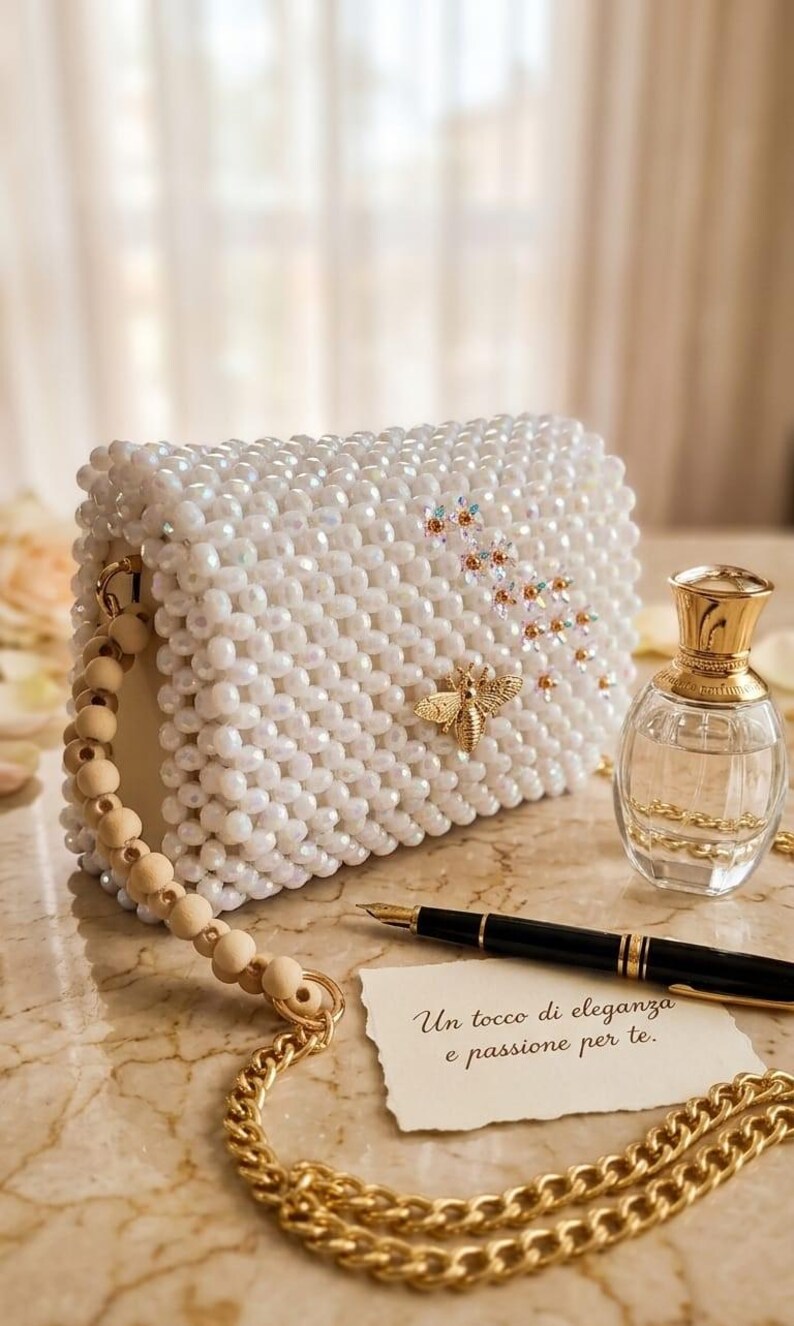 Peut inclure: Un sac &agrave; main blanc perl&eacute; avec une abeille dor&eacute;e et des accents floraux. Le sac a une poign&eacute;e en perles de bois et une bandouli&egrave;re en cha&icirc;ne dor&eacute;e. Un flacon de parfum et un stylo sont &agrave; c&ocirc;t&eacute; du sac. Le texte sur la carte dit "Un tocco di eleganza e passione per te."