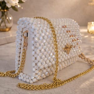Peut inclure: Sac &agrave; main blanc perl&eacute; avec une bandouli&egrave;re en cha&icirc;ne dor&eacute;e. Le sac rectangulaire pr&eacute;sente un embl&egrave;me d'abeille dor&eacute; et de petits accents dor&eacute;s. Le sac est orn&eacute; de perles blanches et poss&egrave;de une bandouli&egrave;re en cha&icirc;ne dor&eacute;e.