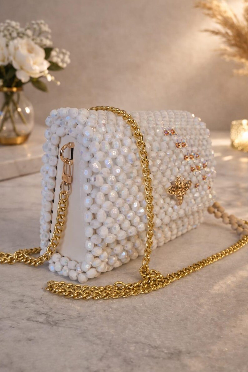 Peut inclure: Sac &agrave; main blanc perl&eacute; avec une bandouli&egrave;re en cha&icirc;ne dor&eacute;e. Le sac rectangulaire pr&eacute;sente un embl&egrave;me d'abeille dor&eacute; et de petits accents dor&eacute;s. Le sac est orn&eacute; de perles blanches et poss&egrave;de une bandouli&egrave;re en cha&icirc;ne dor&eacute;e.
