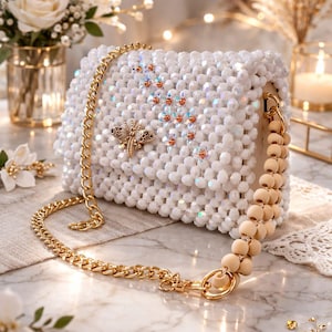 Peut inclure: Un sac &agrave; main blanc perl&eacute; avec une bandouli&egrave;re en cha&icirc;ne dor&eacute;e et une poign&eacute;e en perles de bois. Le sac pr&eacute;sente un embl&egrave;me d'abeille dor&eacute; et est orn&eacute; de perles iris&eacute;es. Le sac est pos&eacute; sur une surface en marbre.