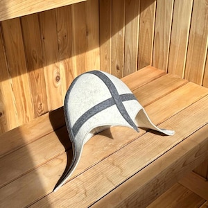 Puede incluir: Un gorro de sauna gris claro con un diseño de cruz gris oscuro. El gorro tiene orejeras y está hecho de fieltro. Está apoyado en un banco de madera dentro de una sauna.