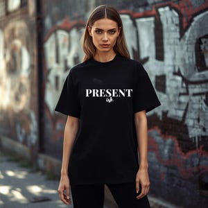 May include: A black short-sleeved t-shirt featuring the word PRESENT in a bold, white, capitalized sans-serif font, with the word ish written below it in a smaller, white, cursive script font. The garment has a crew neckline and a relaxed fit.