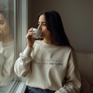Puede incluir: Una sudadera color crema con el texto "I used to pray for quiet... NOW I MISS THE NOISE" en negro. La sudadera tiene mangas largas y cuello redondo. Una persona sostiene una taza blanca.