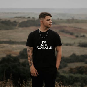 K&ouml;nnte beinhalten: Schwarzes T-Shirt mit dem wei&szlig;en Text "I'M NOT AVAILABLE". Das Shirt ist kurz&auml;rmlig und wird von einer Person getragen. Die Person tr&auml;gt eine silberne Halskette. Der Hintergrund ist eine verschwommene Szene im Freien.