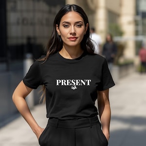 May include: A black short-sleeved t-shirt featuring the white text "PRESENT" in a bold serif font with the word "ish" written in a smaller, cursive script directly underneath. The garment has a crew neckline and a standard fit.