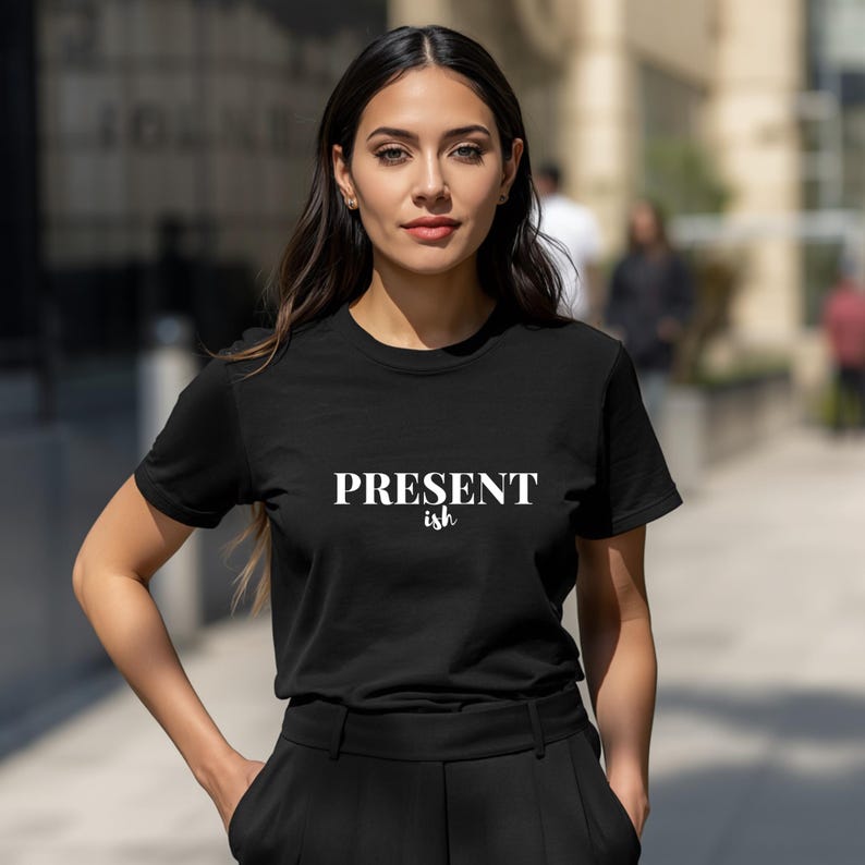 May include: A black short-sleeved t-shirt featuring the white text "PRESENT" in a bold serif font with the word "ish" written in a smaller, cursive script directly underneath. The garment has a crew neckline and a standard fit.