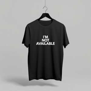K&ouml;nnte beinhalten: Schwarzes T-Shirt mit Rundhalsausschnitt und dem wei&szlig;en Text "I'M NOT AVAILABLE" auf der Vorderseite. Das T-Shirt h&auml;ngt an einem schwarzen Kleiderb&uuml;gel vor einem neutralen Hintergrund. Das kurz&auml;rmelige Shirt ist aus weichem Material.