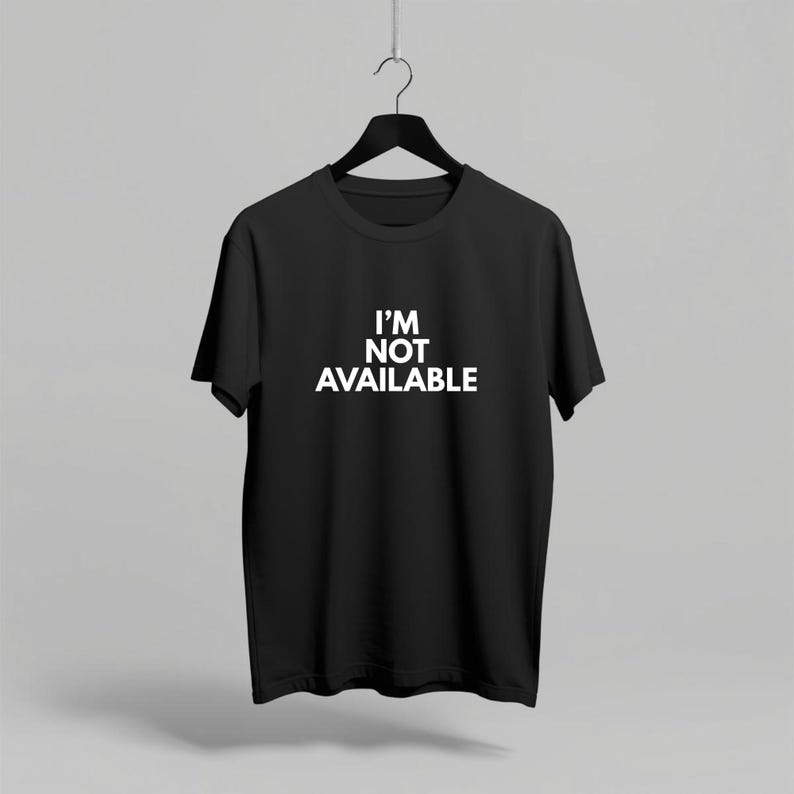 K&ouml;nnte beinhalten: Schwarzes T-Shirt mit Rundhalsausschnitt und dem wei&szlig;en Text "I'M NOT AVAILABLE" auf der Vorderseite. Das T-Shirt h&auml;ngt an einem schwarzen Kleiderb&uuml;gel vor einem neutralen Hintergrund. Das kurz&auml;rmelige Shirt ist aus weichem Material.
