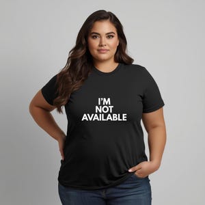 K&ouml;nnte beinhalten: Schwarzes T-Shirt mit Rundhalsausschnitt und dem wei&szlig;en Text "I'M NOT AVAILABLE". Das Shirt wird mit einer blauen Jeans kombiniert. Das Model steht vor einem grauen Hintergrund.