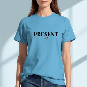 May include: A light blue short-sleeve t-shirt featuring the text "PRESENT ish" printed in black across the chest. The word "PRESENT" is in a bold, capitalized serif font, while the word "ish" is in a smaller, cursive script font.