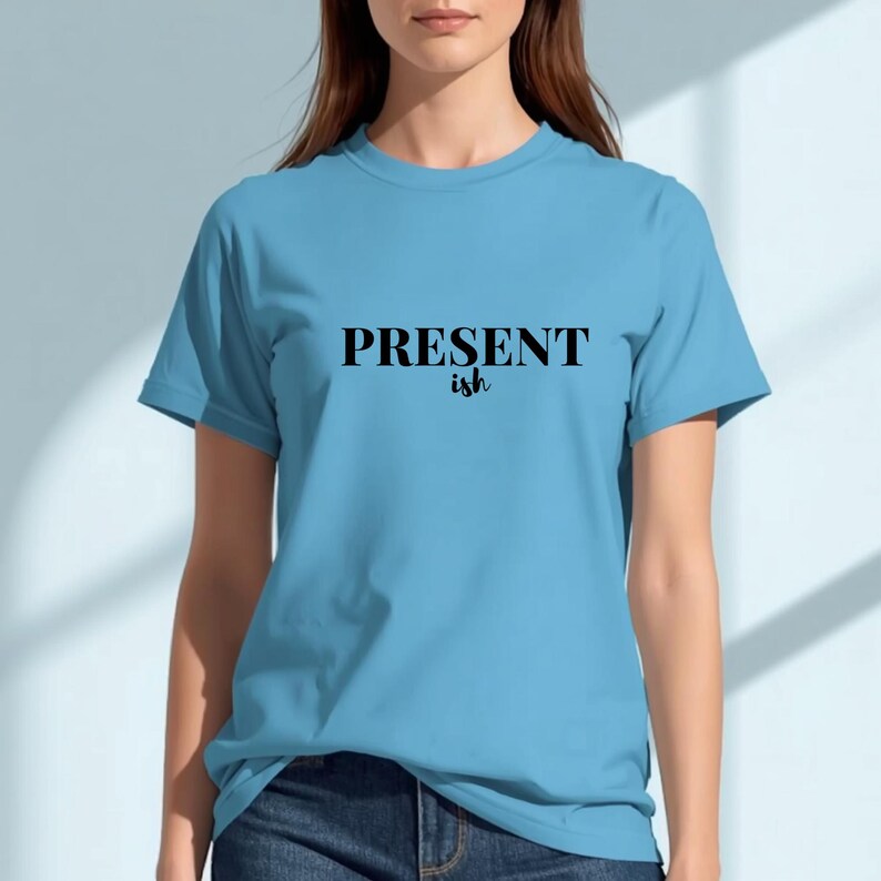 May include: A light blue short-sleeve t-shirt featuring the text "PRESENT ish" printed in black across the chest. The word "PRESENT" is in a bold, capitalized serif font, while the word "ish" is in a smaller, cursive script font.
