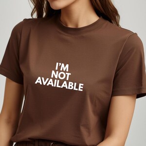 K&ouml;nnte beinhalten: Ein braunes T-Shirt mit dem wei&szlig;en Text "I'M NOT AVAILABLE". Das kurz&auml;rmlige Oberteil hat einen Rundhalsausschnitt und ist aus einem weichen Material gefertigt. Es ist ein l&auml;ssiges Kleidungsst&uuml;ck.