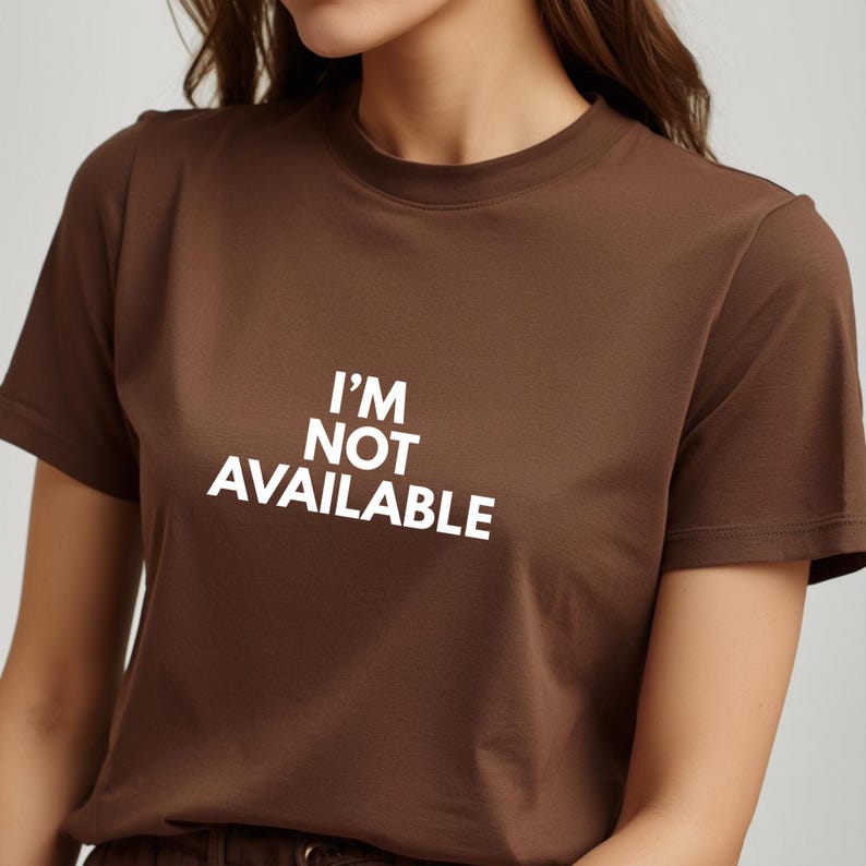 K&ouml;nnte beinhalten: Ein braunes T-Shirt mit dem wei&szlig;en Text "I'M NOT AVAILABLE". Das kurz&auml;rmlige Oberteil hat einen Rundhalsausschnitt und ist aus einem weichen Material gefertigt. Es ist ein l&auml;ssiges Kleidungsst&uuml;ck.