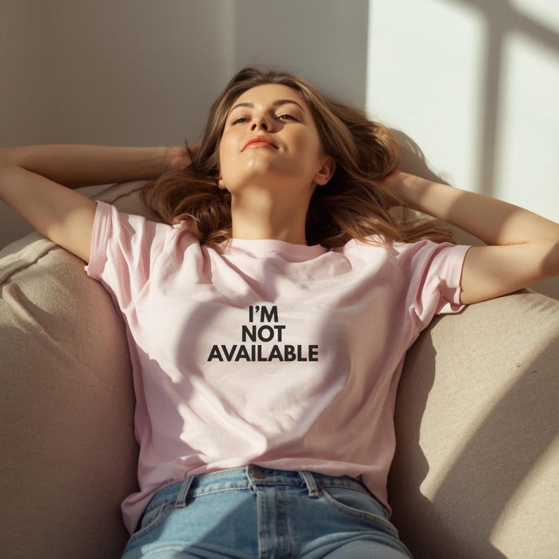 K&ouml;nnte beinhalten: Hellrosa T-Shirt mit dem schwarzen Text "I'M NOT AVAILABLE". Die Person tr&auml;gt eine blaue Jeans. Die Person sitzt auf einem beigen Sofa.