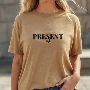 May include: A tan short-sleeved t-shirt featuring the word PRESENT in a bold, black, sans-serif font, with the word ish written underneath in a smaller, black, cursive script. The garment has a crew neckline and a standard fit.