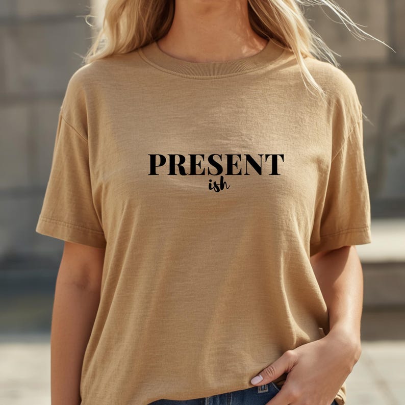 May include: A tan short-sleeved t-shirt featuring the word PRESENT in a bold, black, sans-serif font, with the word ish written underneath in a smaller, black, cursive script. The garment has a crew neckline and a standard fit.