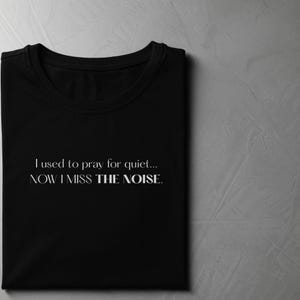 May include: Black t-shirt with the text "I used to pray for quiet... NOW I MISS THE NOISE." in a white, elegant font. The shirt is folded neatly, showcasing the design on a neutral background.