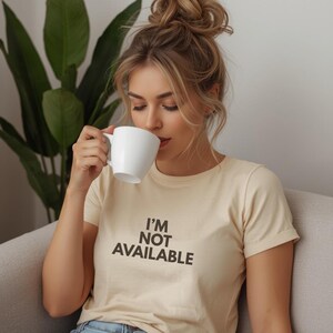K&ouml;nnte beinhalten: Eine Frau tr&auml;gt ein cremefarbenes T-Shirt mit dem schwarzen Text "I'M NOT AVAILABLE". Sie trinkt aus einer wei&szlig;en Tasse. Die Frau hat blonde Haare und sitzt auf einem grauen Sofa. Im Hintergrund ist eine gr&uuml;ne Pflanze zu sehen.