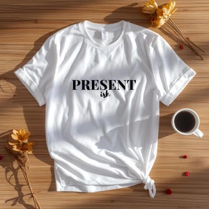 May include: A white short-sleeved t-shirt featuring the black text "PRESENT ish" printed on the chest. The shirt has a crew neckline and a knotted hem detail on the lower right side, showcasing a clean design with simple typography and fabric folds.